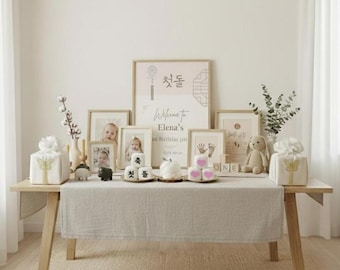 May include: A light wood table is set for a first birthday party. The table is decorated with framed photos, gifts, a stuffed bunny, and a sign that reads "Welcome to Elena's 1st Birthday party". The table is covered with a light gray linen table runner.