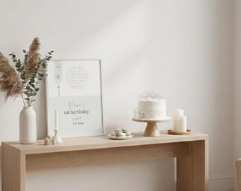 May include: A light wood console table displays a framed print, a white cake on a wooden stand, candles, and a vase with dried plants. The print reads "Mama's 1st Birthday" with a date. Small desserts are on a plate.