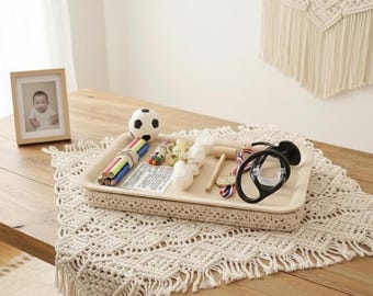 May include: A beige tray with a woven border holds a soccer ball, colored pencils, and small toys. A framed photo and a macrame wall hanging are also visible. The tray sits on a wooden table with a macrame runner.