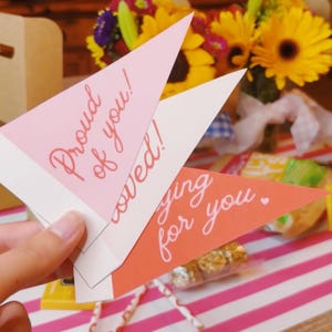 May include: Three triangular cards with messages, held in hand, against a backdrop of flowers and a striped table. The cards are pink and orange, with the phrases "Proud of you!", "Loved!", and "Caring for you."