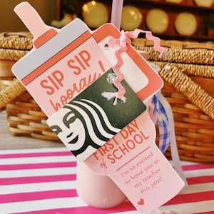 May include: A pink tumbler with a straw and a gift card holder. The holder is shaped like a bottle and has the words "SIP SIP hooray!" and "FIRST DAY of SCHOOL" printed on it. A gift card is attached with a pink ribbon.