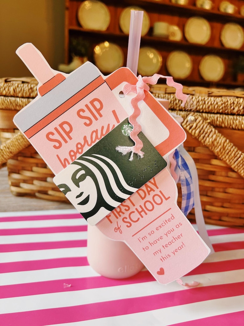 May include: A pink tumbler with a straw and a gift card holder. The holder is shaped like a bottle and has the words "SIP SIP hooray!" and "FIRST DAY of SCHOOL" printed on it. A gift card is attached with a pink ribbon.