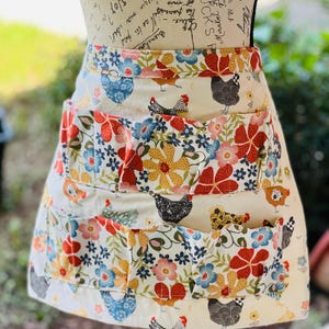 May include: A cream-colored apron with a floral and chicken pattern. The apron features three pockets and is adorned with red, yellow, blue, and orange flowers, along with various chicken illustrations. The apron is designed for kitchen use.