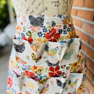 May include: An egg-gathering apron with multiple pockets, featuring a colorful floral and chicken print. The apron has a white background with red, yellow, and blue flowers, and various chicken illustrations. The apron is designed for practical use.