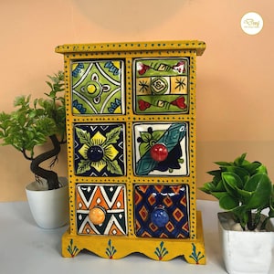 May include: A yellow wooden storage chest with six drawers, each featuring unique hand-painted ceramic fronts with colorful geometric and floral patterns. The frame is decorated with small blue dots, and each drawer has a round knob in a solid color.
