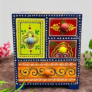 Op de afbeelding: Een kleine, vierkante, handgeschilderde houten ladekast. De kast is donkerblauw geschilderd met vier lades, elk met een andere kleur en een ander ontwerp. De lades hebben oranje, rode en groene achtergronden met decoratieve knoppen.