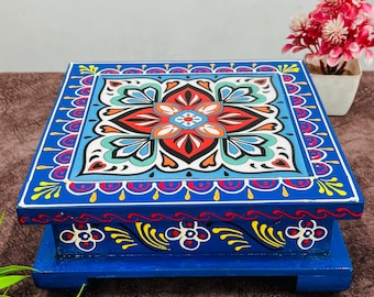 May include: A square, blue wooden decorative stand with a detailed floral design on the top surface. The floral design features red, orange, white, and blue colours. The stand has a decorative border and is supported by a base.