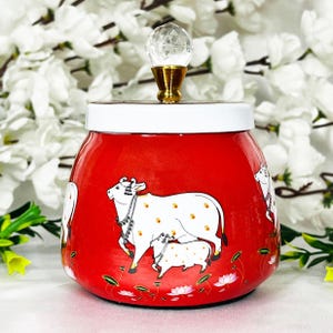 May include: Red ceramic jar with a white lid and a crystal knob. The jar features a painted design of two white cows with black spots on a red background.
