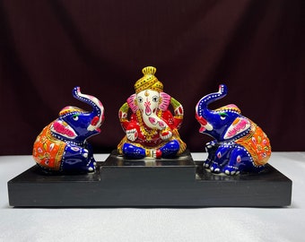 May include: A decorative set featuring a central figure of Ganesha, flanked by two elephants. The figures are vibrantly painted with blue, orange, pink, and gold details. The set is displayed on a black wooden base.