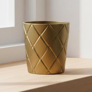 Puede incluir: Una maceta cilíndrica de color dorado mate que presenta un patrón geométrico de diamantes en relieve en toda su superficie exterior. El recipiente tiene una amplia abertura circular en la parte superior y se estrecha hacia la base.