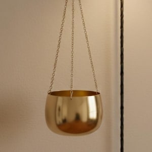 Puede incluir: Una maceta colgante de metal dorado con forma de cuenco redondeado, suspendida por tres cadenas doradas finas conectadas a un gancho en forma de S. La maceta está situada junto a una lámpara de pared negra con cable y un cuadro abstracto.