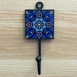 Könnte beinhalten: Ein dekorativer Wandhaken mit einer quadratischen, blauen Fliese mit einem floralen Design in Gelb und Weiß. Die Fliese ist in schwarzem Metall eingefasst und hat einen schwarzen Haken. Der Haken ist an einem schwarzen Metallring zum Aufhängen befestigt.