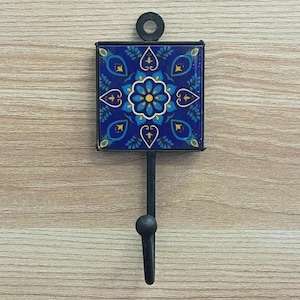 Könnte beinhalten: Ein dekorativer Wandhaken mit einer quadratischen, blauen Keramikfliese mit einem Blumendesign in Blau- und Gelbtönen. Die Fliese ist in schwarzem Metall eingefasst und hat einen schwarzen Haken darunter. Oben befindet sich ein schwarzer Ring zum Aufhängen.