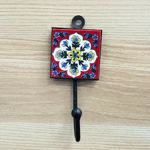 Könnte beinhalten: Ein dekorativer Wandhaken mit einer quadratischen, roten Fliese mit einem floralen Design in Gelb, Blau und Weiß. Die Fliese ist in schwarzem Metall gerahmt, mit einem schwarzen Haken und einem Befestigungsring. Der Haken ist ca. 15 cm lang.