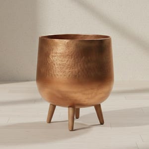 Puede incluir: Una maceta de color cobre con una textura martillada. La maceta redonda se asienta sobre tres patas de madera. El interior de la maceta es oscuro y el exterior tiene un brillo metálico. La maceta está diseñada para uso en interiores o exteriores.