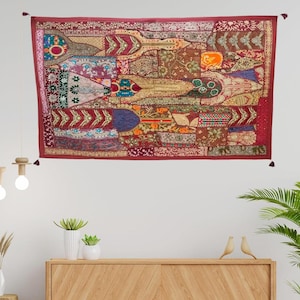 Puede incluir: Un tapiz de pared rectangular de patchwork en tonos rojos, marrones y dorados. El tapiz presenta una variedad de patrones y texturas, con un borde y borlas de color rojo oscuro. Se exhibe en una pared blanca.