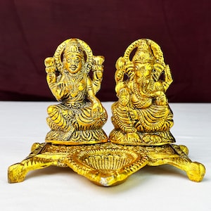 May include: Gold metal diya with two deities, Lakshmi and Ganesha, sitting on a platform. The diya has three small bowls for oil and wicks.