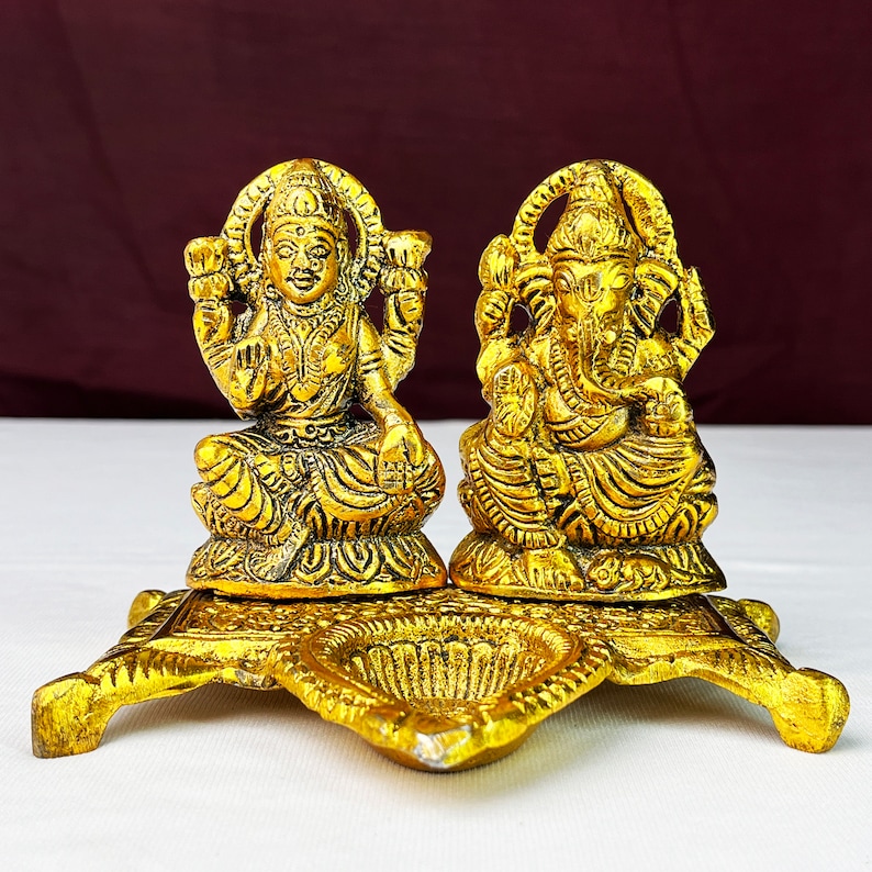 May include: Gold metal diya with two deities, Lakshmi and Ganesha, sitting on a platform. The diya has three small bowls for oil and wicks.