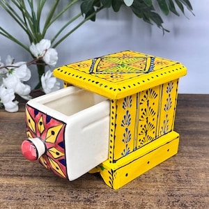 May include: A yellow wooden storage box with a single pull-out ceramic drawer. The exterior features hand-painted blue floral patterns and black dots. The drawer front displays a red, yellow, and blue geometric star design with a round red knob.