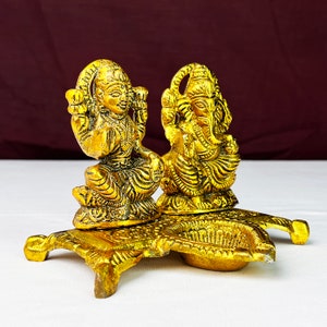May include: Gold-toned metal diya with two figures of Hindu deities, Lakshmi and Ganesha, sitting on a decorative base.