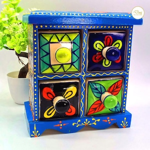 May include: A blue, hand-painted wooden storage chest with four square drawers. Each drawer features a unique floral design in vibrant colors like green, red, and yellow. The chest has a decorative trim and knobs.