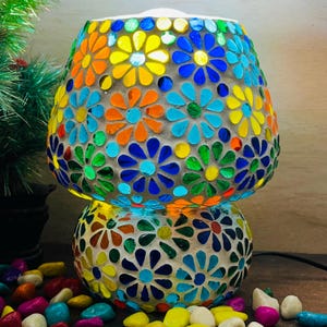 Puede incluir: Una lámpara decorativa de mosaico con un diseño floral. La lámpara presenta una base y una pantalla de dos niveles, adornadas con vibrantes baldosas de vidrio en tonos azules, naranjas, amarillos y verdes. Un elemento decorativo para el hogar.