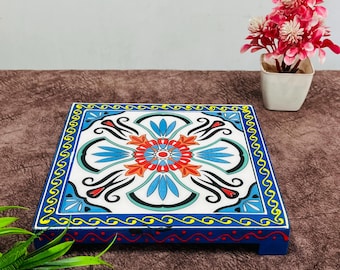 May include: A square, wooden decorative tray with a vibrant floral design. The tray features a white background with a central orange and blue flower motif, surrounded by blue and black accents. The edges are a bright blue with yellow and red detailing.