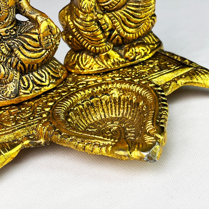 May include: Gold metal diya with two small figures sitting on top. The diya has a decorative design and is used for burning oil lamps.