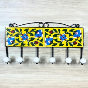 Puede incluir: Un perchero de pared de azulejos amarillos con seis ganchos de cerámica blanca. Los azulejos presentan un patrón floral azul y verde.