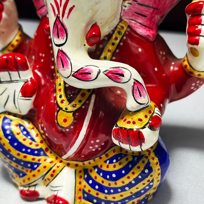 May include: A red, white and gold painted ceramic figurine of Ganesha, the Hindu god of wisdom and prosperity. The figurine is sitting with its trunk raised and its hands in a gesture of blessing. The figurine is decorated with intricate details, including a floral pattern on the trunk and a necklace of beads around the neck.