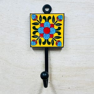 Könnte beinhalten: Ein schwarzer Metallhaken mit einer gelben Keramikfliese mit einem blauen und roten Blumendesign. Die Fliese hat einen schwarzen Rand.