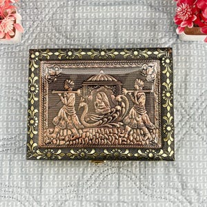 Puede incluir: Una caja de madera decorativa con un diseño de cobre de una procesión que lleva un palanquín. La caja tiene un borde dorado con motivos florales.