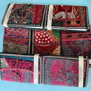 Könnte beinhalten: Eine Sammlung handgefertigter Papiernotizbücher mit farbenfrohen Patchwork-Einbänden. Die Einbände weisen eine Vielzahl von Texturen auf, darunter Stickereien und Perlen, in Rot-, Rosa- und Türkistönen. Jedes Notizbuch ist mit einem weißen Band und Bindfaden gebunden.