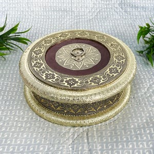 Puede incluir: Una caja redonda de metal de color dorado con intrincados diseños florales. La tapa tiene un centro marrón con un diseño de estrella dorada y una pequeña manija de metal.