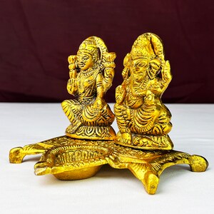 May include: Gold-toned metal diya with two deities, Lakshmi and Ganesha, sitting on a decorative base. The diya has two small bowls for oil and wicks.