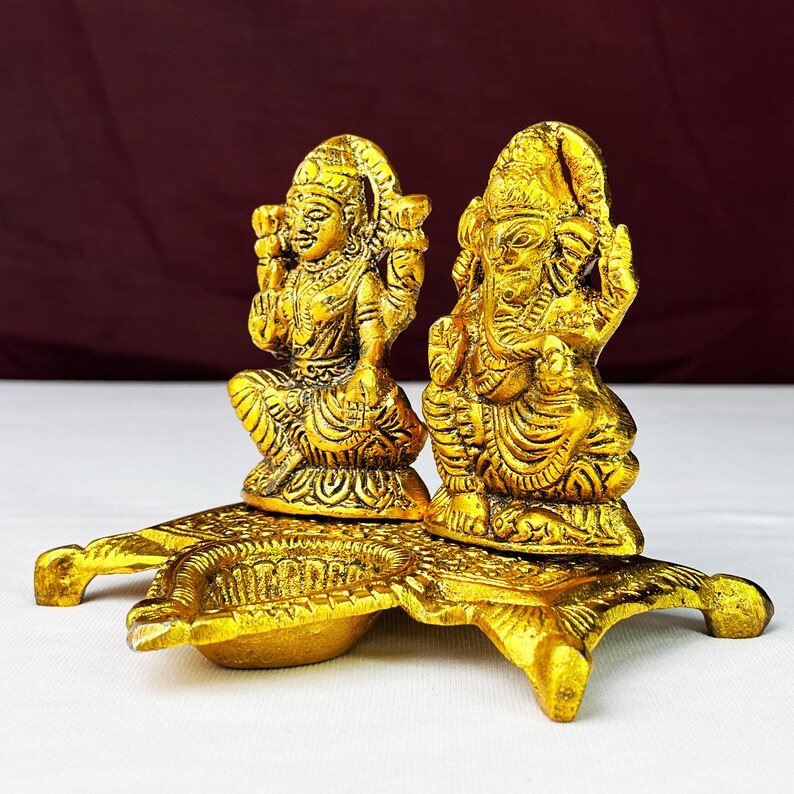 May include: Gold-toned metal diya with two deities, Lakshmi and Ganesha, sitting on a decorative base. The diya has two small bowls for oil and wicks.