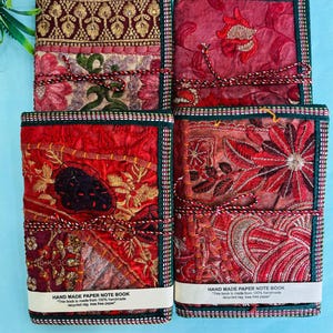 Könnte beinhalten: Vier handgefertigte Papiernotizbücher mit farbenfrohen, bestickten Stoffeinbänden. Die Einbände zeigen rote, rosa und goldene Blumenmuster mit einem grünen Rand. Jedes Notizbuch ist mit einer dekorativen Kordel gebunden und hat ein weißes Etikett.