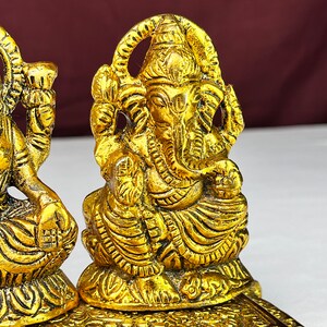 May include: Two gold-colored metal statues of Hindu deities, one of Ganesha and one of Lakshmi, sitting on a decorative base.