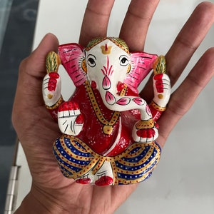 May include: A colourful, hand-painted figurine of the Hindu deity Ganesha. The figurine is made of metal and depicts Ganesha sitting with his trunk raised and holding a sweet in his hand. He is adorned with a red and blue patterned dhoti and a gold necklace.