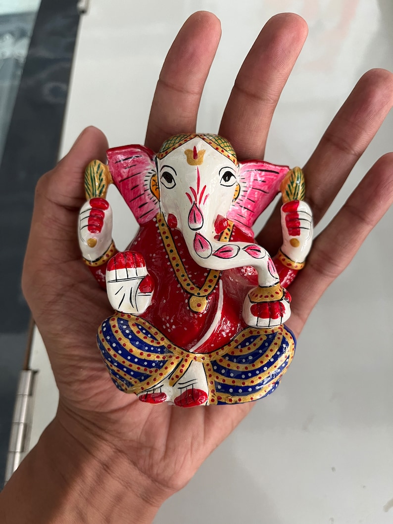 May include: A colourful, hand-painted figurine of the Hindu deity Ganesha. The figurine is made of metal and depicts Ganesha sitting with his trunk raised and holding a sweet in his hand. He is adorned with a red and blue patterned dhoti and a gold necklace.