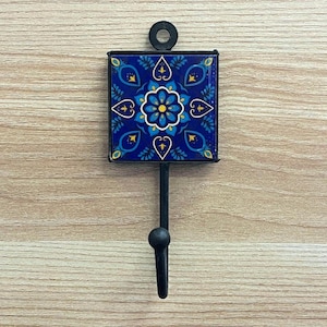 Könnte beinhalten: Ein dekorativer Wandhaken mit einer quadratischen, blauen Keramikfliese mit einem Blumendesign in Blau- und Gelbtönen. Die Fliese ist in schwarzem Metall gerahmt, mit einem schwarzen Haken und einem kreisförmigen Montage ring.