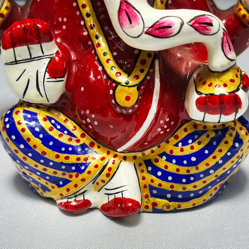 May include: A red, white and gold painted figurine of Ganesha, the Hindu god of wisdom and prosperity. The figurine is sitting with its legs crossed and has a trunk, large ears, and a single tusk. The figurine is decorated with intricate details and colourful patterns.
