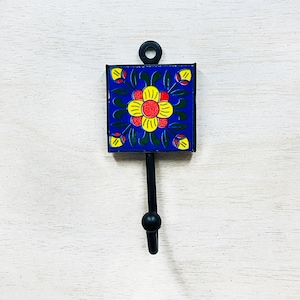 Könnte beinhalten: Ein schwarzer Metallhaken mit einem blauen und gelben Fliesenmuster mit Blumen. Die Fliese zeigt eine rote Blume mit gelben Blütenblättern und grünen Blättern.