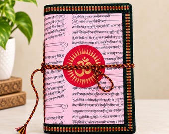 Könnte beinhalten: Ein rechteckiges Notizbuch mit hellrosa Einband, schwarzem Sanskrit-Text und einem zentralen roten kreisförmigen Emblem mit goldenem Om-Symbol. Der Einband ist mit dunkelgrünem Stoff mit rot-gelbem Muster eingefasst und mit einer Kordel gebunden.