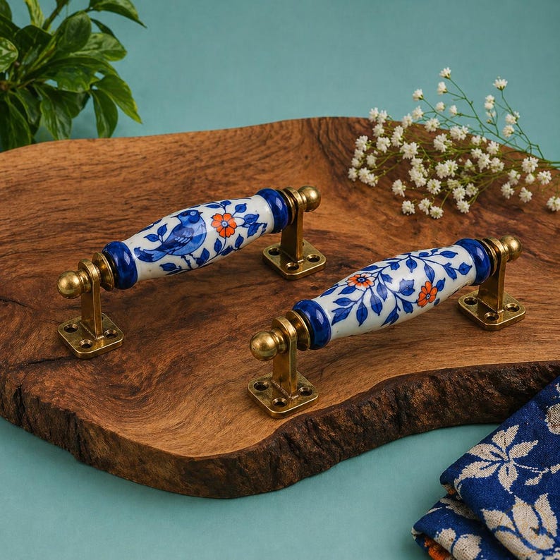 May include: A pair of decorative cabinet pulls. The white ceramic handles have blue bird and floral patterns. They have brass-coloured metal bases and accents. The pulls are displayed on a wooden chopping board.