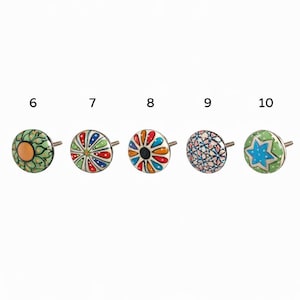 Op de afbeelding: Vijf decoratieve keramische kastknoppen met metalen stelen. De knoppen hebben kleurrijke ontwerpen, waaronder bloempatronen, sterren en geometrische vormen. Kleuren zijn onder meer groen, oranje, blauw, rood en wit. Deze knoppen zijn ideaal om een unieke touch aan meubels toe te voegen.