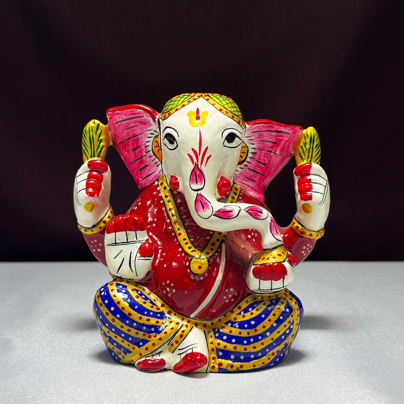 May include: A colourful hand-painted metal figurine of the Hindu deity Ganesha. The figurine is red, pink, white, blue, and yellow. Ganesha is depicted with an elephant head, a single tusk, and four arms. He is sitting with his legs crossed and holding a sweet in one hand.