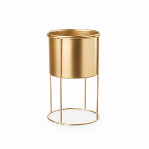 May include: A cylindrical gold-coloured metal planter with a smooth, brushed finish. The container is supported by a thin, open-frame wire stand consisting of four vertical rods connected to a circular base ring, creating a tall, elevated structure.
