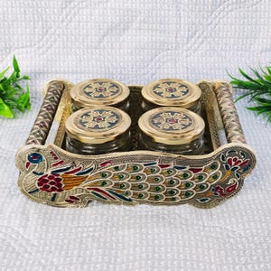 Puede incluir: Una bandeja de metal dorado con un diseño de pavo real y cuatro tarros de vidrio con tapas doradas. Los tarros tienen intrincados diseños florales en las tapas.