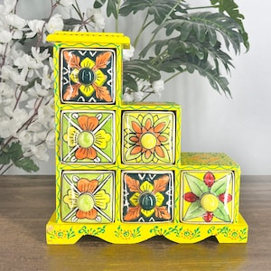 May include: A yellow, tiered wooden storage box with six square drawers. Each drawer is decorated with a floral design in orange, green, and yellow. The box has a decorative base and a small top.